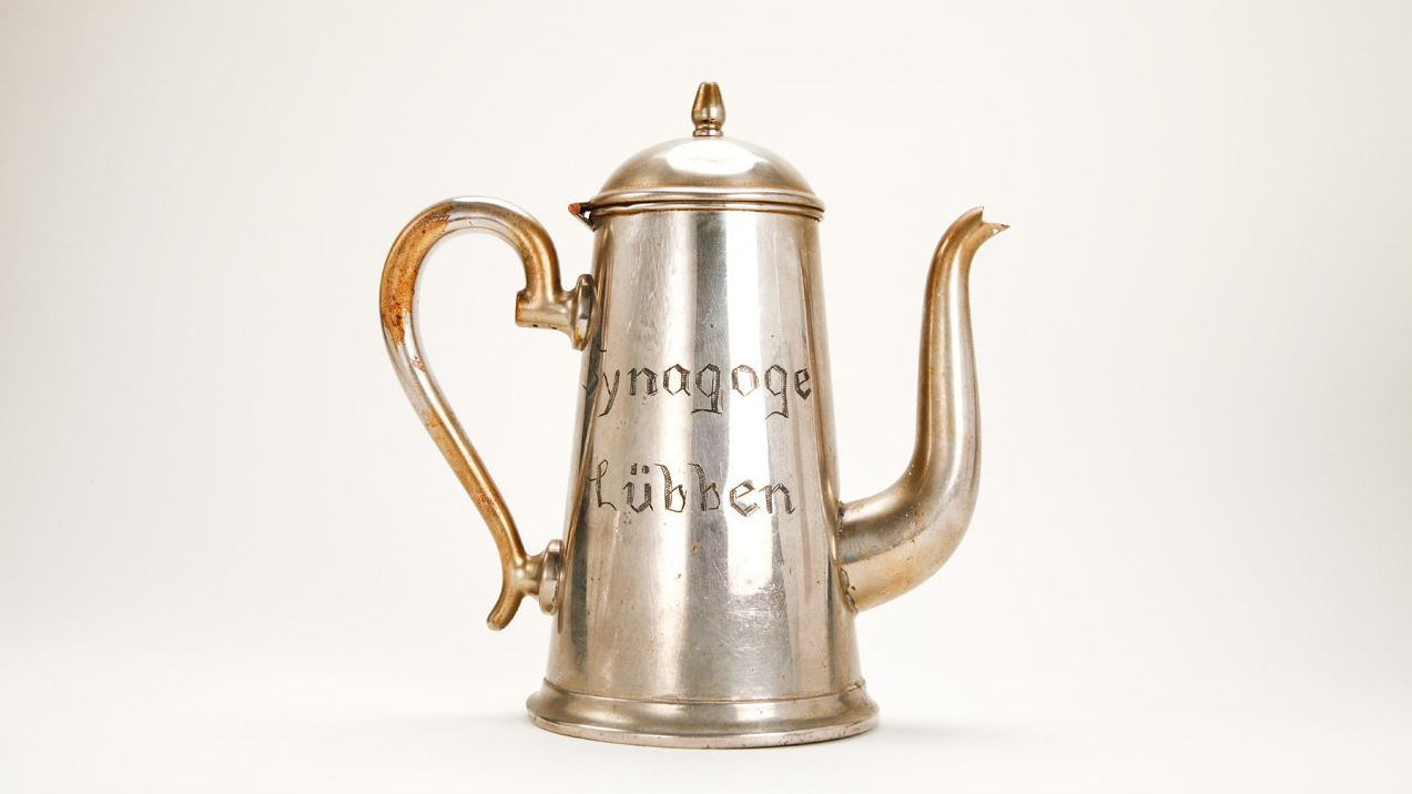 Das Bild zeigt eine versilberte Kanne aus Messing und Zinn die zwei Gravuren trägt: „Synagoge Lübben“ und einen sechseckigen Stern, der aus zwei miteinander verwobenen Dreiecken besteht: ein Davidsstern.