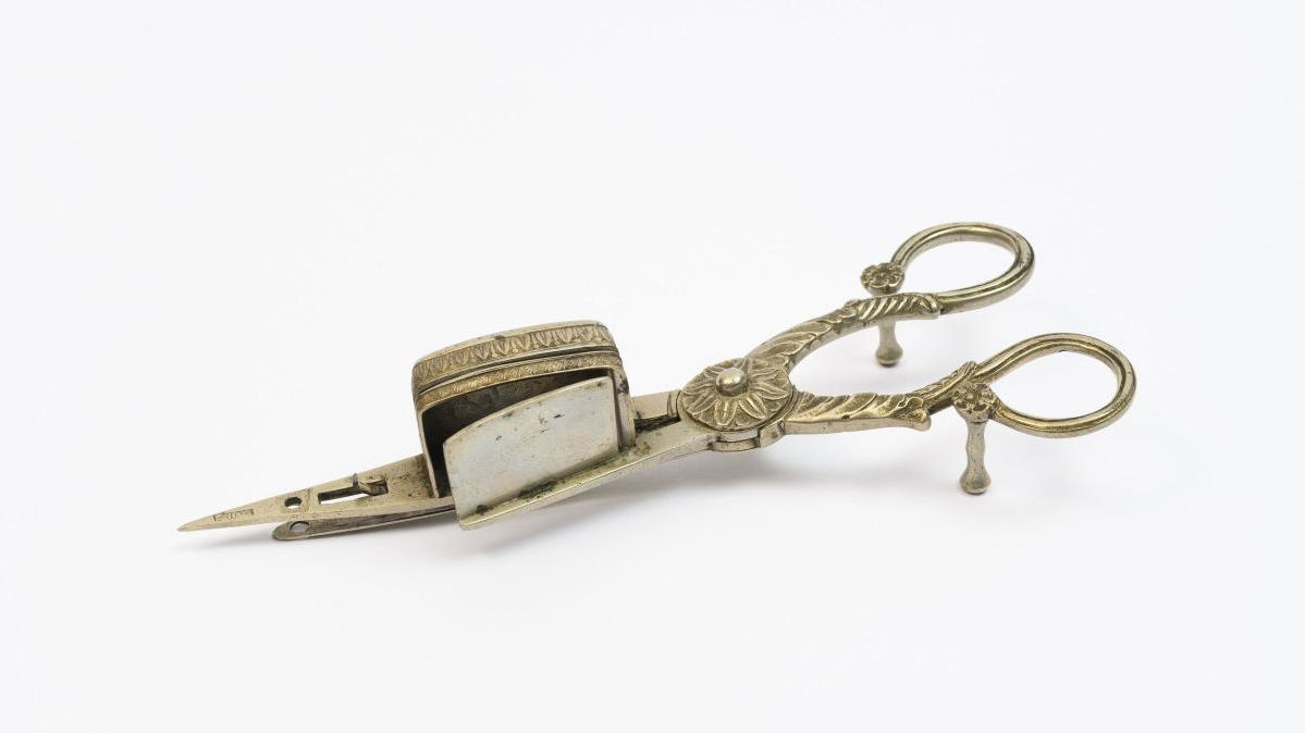 Foto: Eine verzierte Metallschere mit einem kleinen Kästchen an einer der Scherenspitzen und einem Deckel an der Anderen.